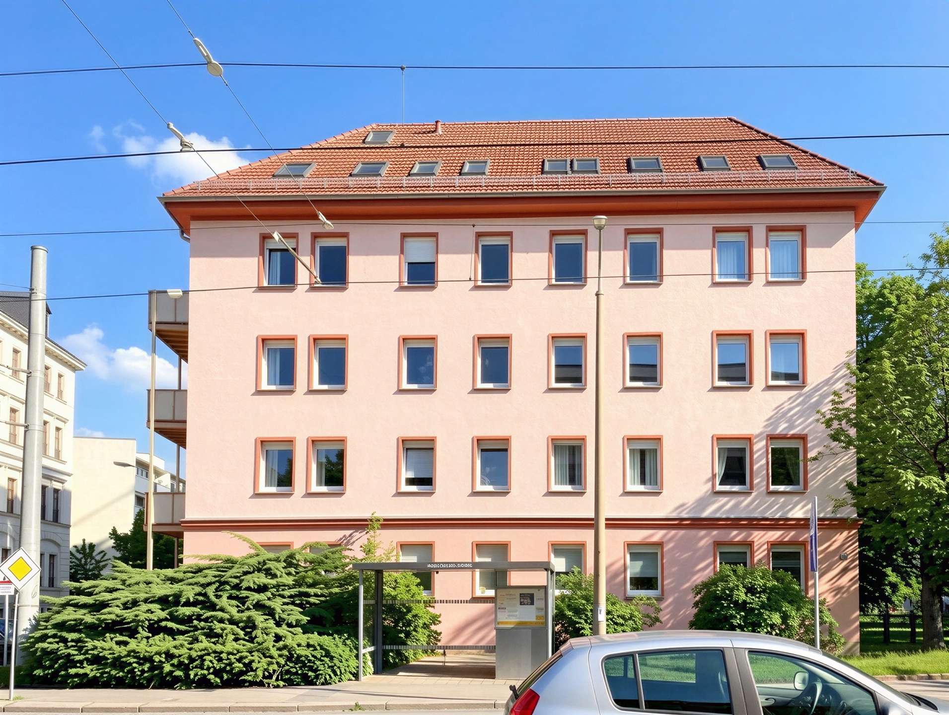 Pink four-story building with a red-tiled roof and rows of rectangular windows beneath a clear blue sky, street-level greenery in front.