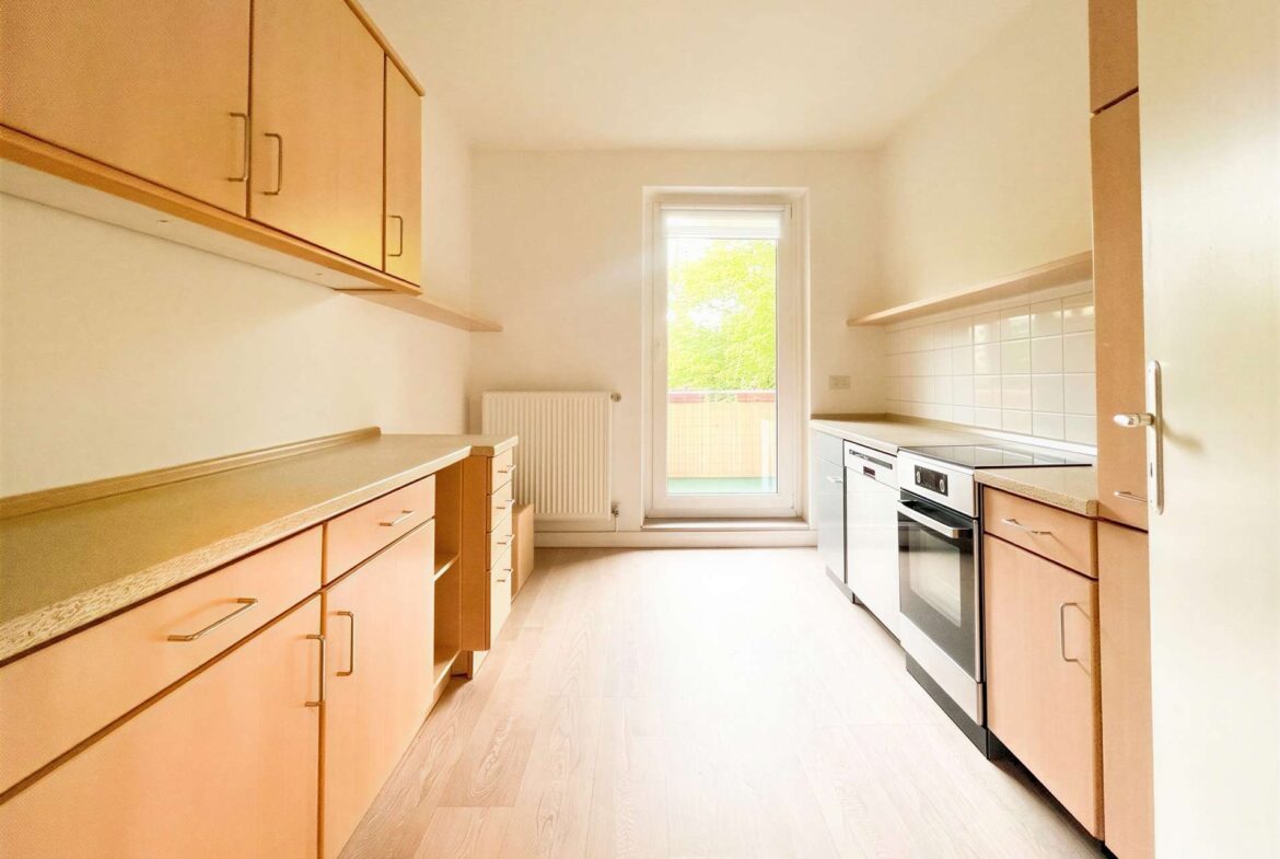 Bright, narrow kitchen with light wood cabinets, beige countertops, and a stainless steel oven along one wall; a window at the far end lets in natural light.