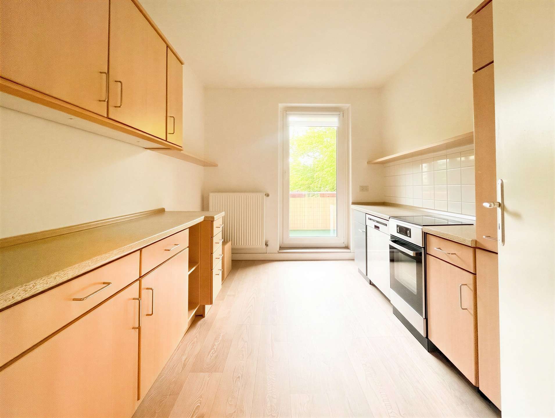 Bright, narrow kitchen with light wood cabinets, beige countertops, and a stainless steel oven along one wall; a window at the far end lets in natural light.
