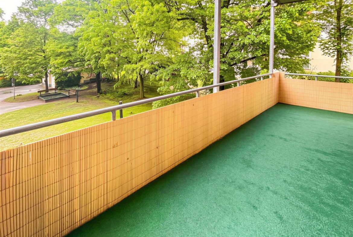 Balcony with green turf and a beige bamboo privacy wall, metal railing, overlooking a park with leafy trees.