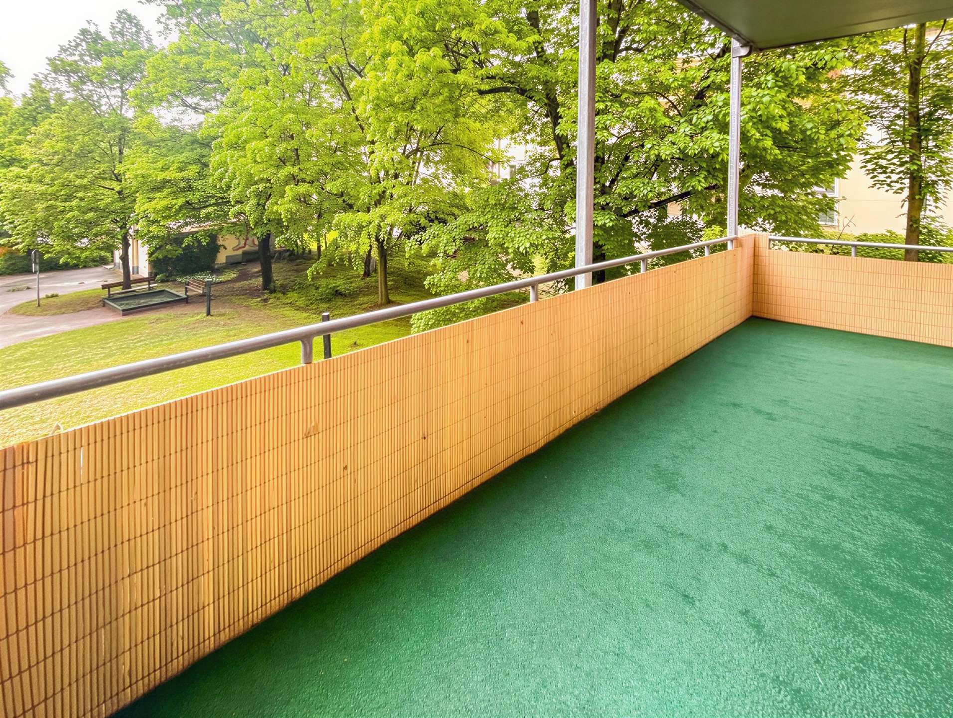 Balcony with green turf and a beige bamboo privacy wall, metal railing, overlooking a park with leafy trees.