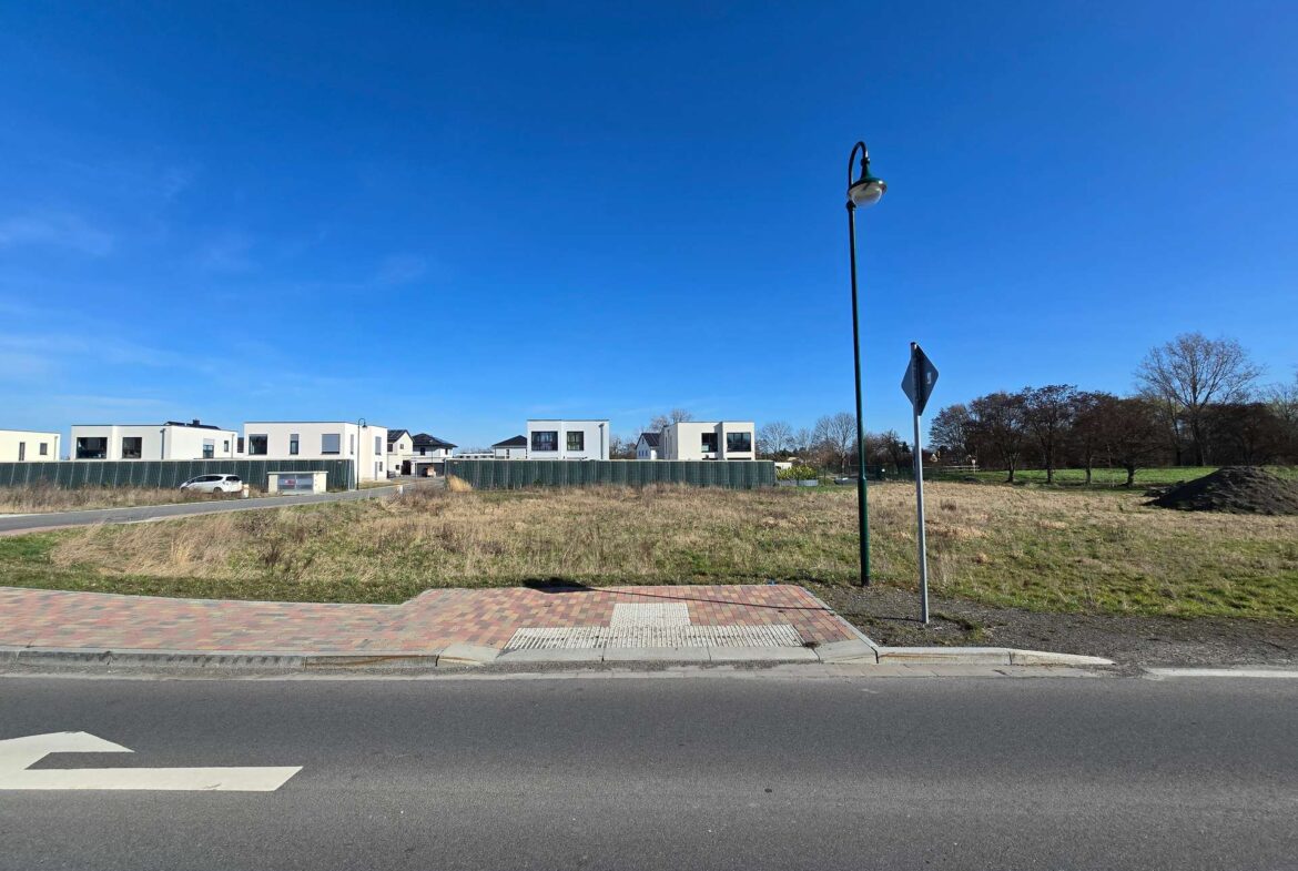 Wide street view of a suburban development: row of white modern houses behind a green construction fence, a white car parked on the left, and a tall street lamp with a curved top in the midground under a clear blue sky.