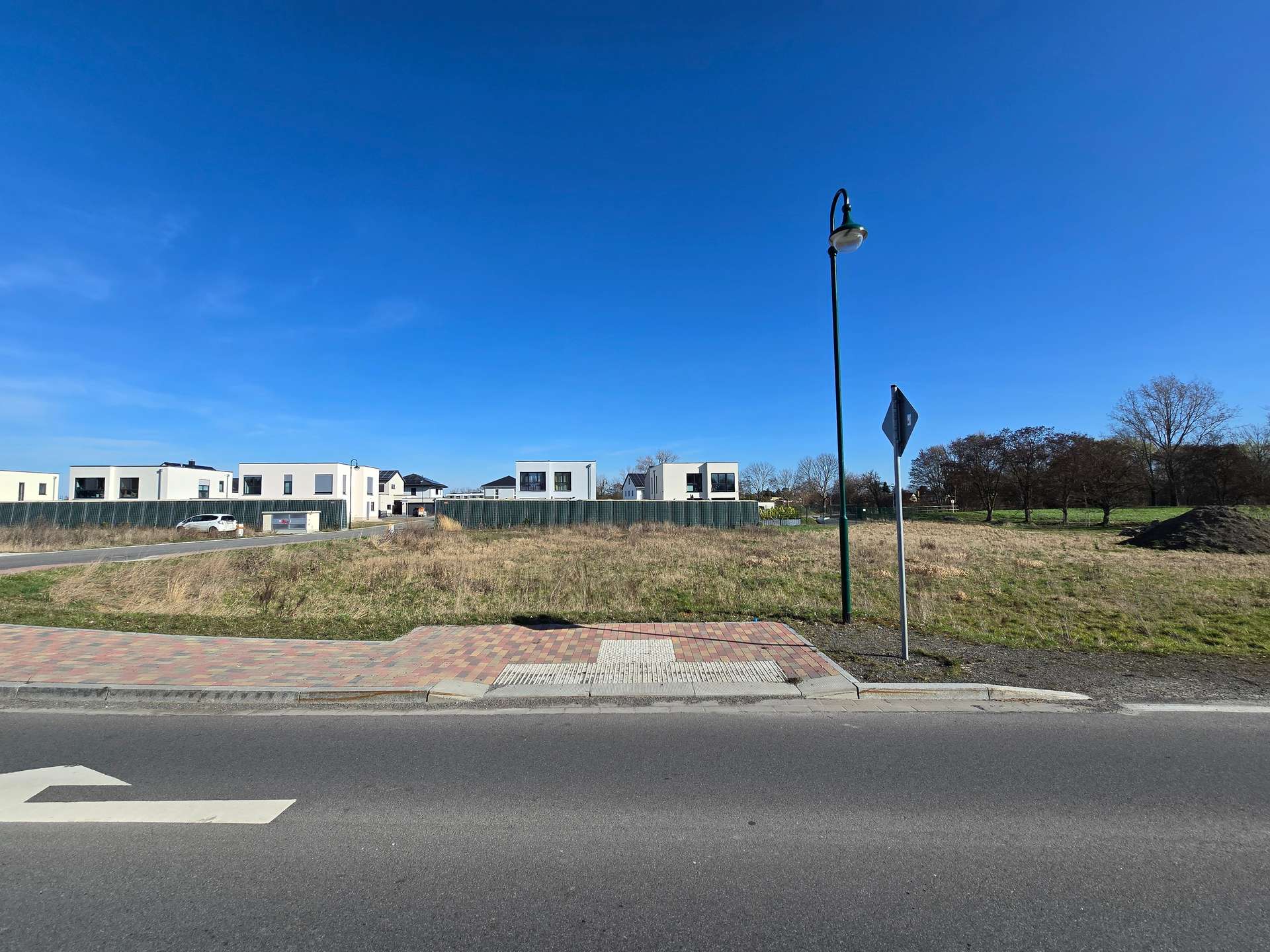 Wide street view of a suburban development: row of white modern houses behind a green construction fence, a white car parked on the left, and a tall street lamp with a curved top in the midground under a clear blue sky.
