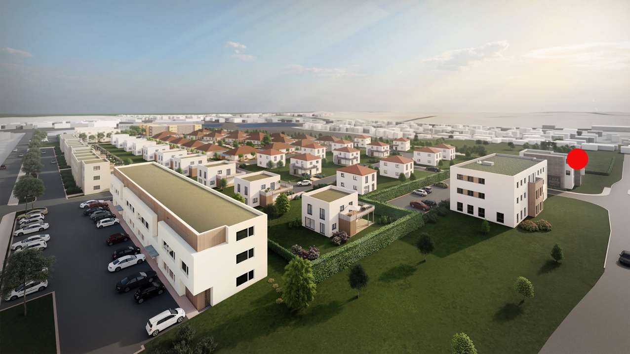 Aerial view of a modern housing development with white low-rise buildings, red-roofed homes, green lawns, and winding roads; a red marker highlights a building on the right.