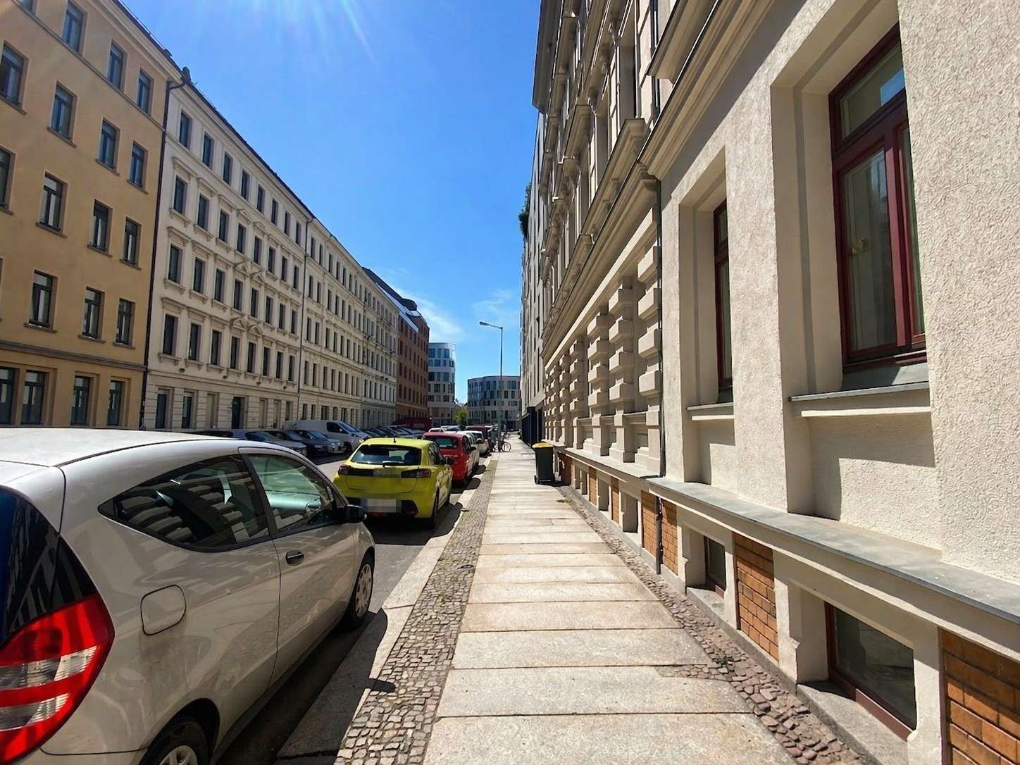 Sunny urban street scene with parked cars along the curb, beige and white buildings on both sides and a bright blue sky.
