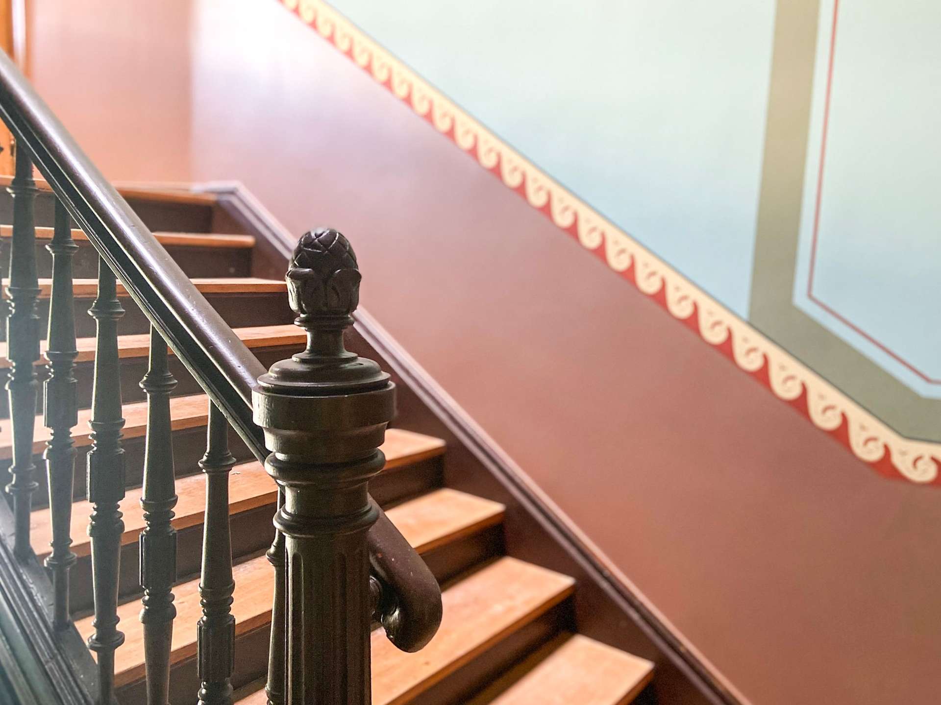 Decorative black iron staircase railing with a carved newel post beside wooden steps and a diagonal patterned border on a pastel wall.