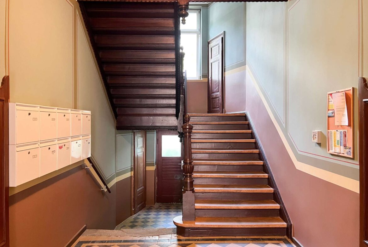 Wooden staircase with a dark banister winding up a vintage hallway, mailboxes on the left and a door at the landing ahead; patterned tiled floor and decorative wall trim.
