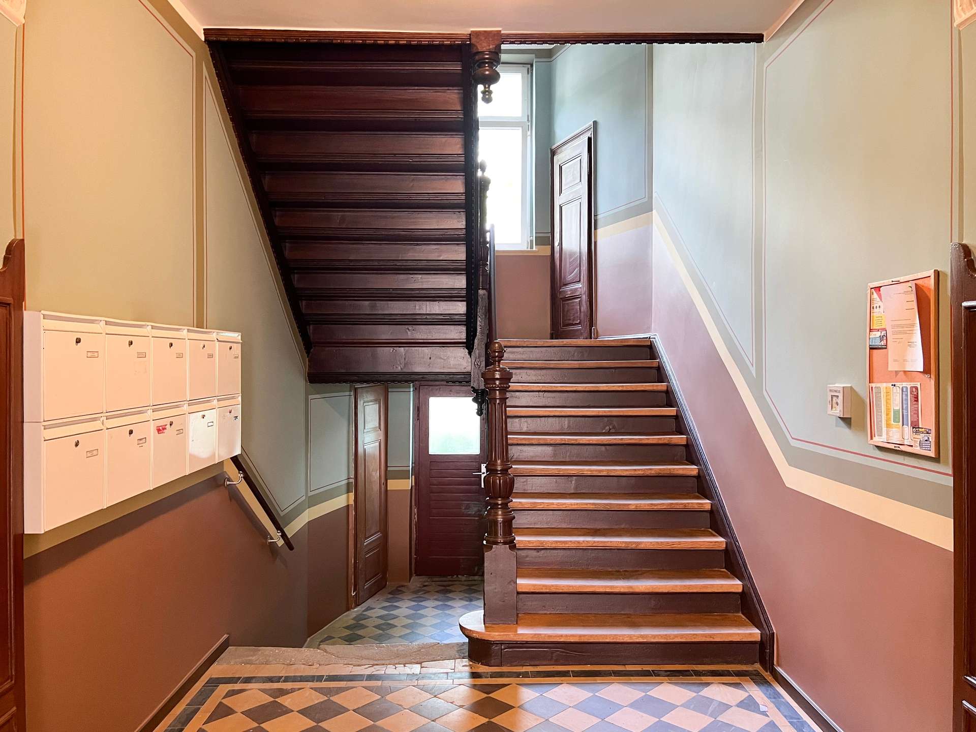 Wooden staircase with a dark banister winding up a vintage hallway, mailboxes on the left and a door at the landing ahead; patterned tiled floor and decorative wall trim.