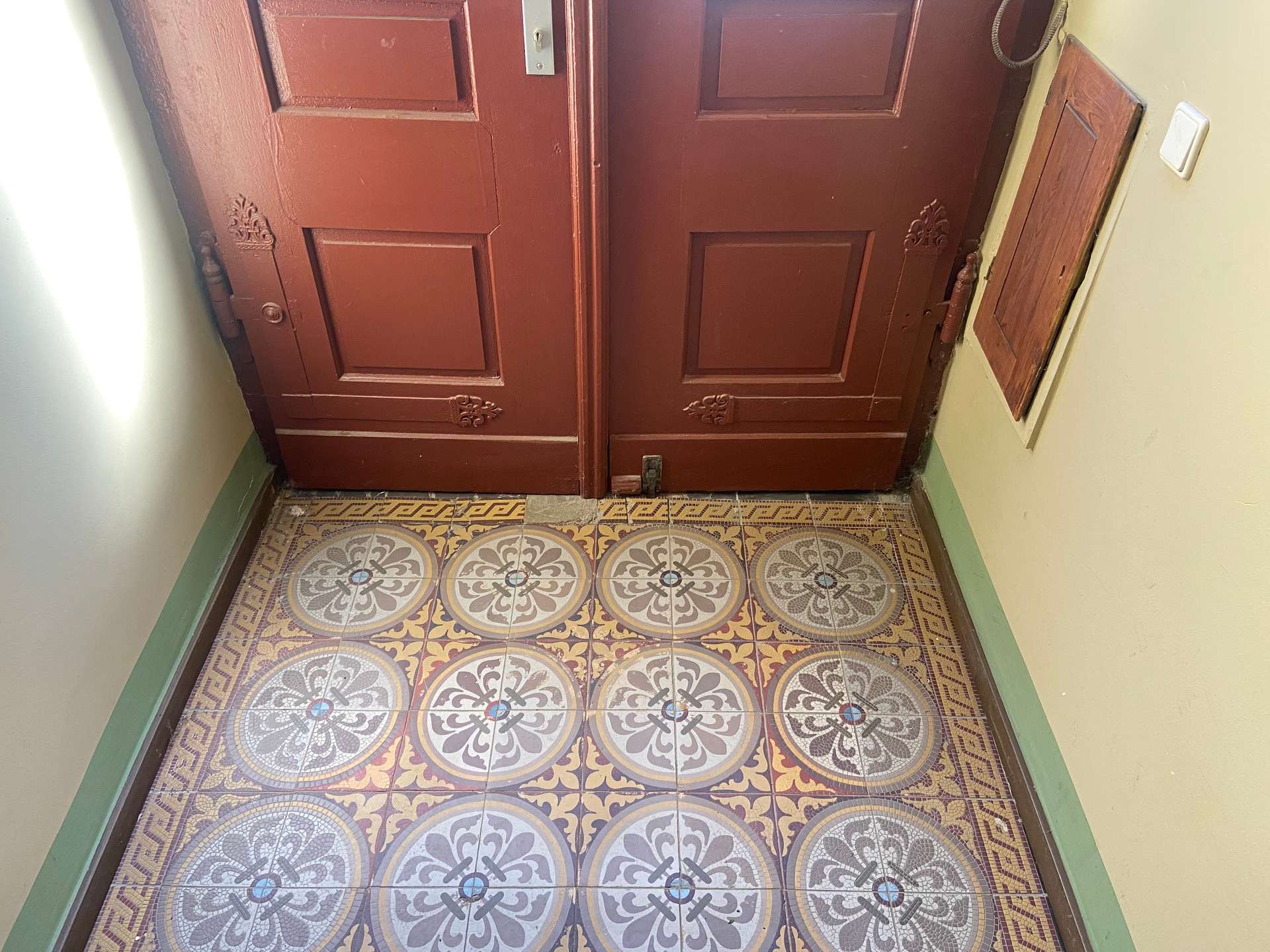 Pair of dark brown wooden doors with decorative panels at the end of a narrow hallway, carved details visible, tiled floor with circular patterns and green baseboards.