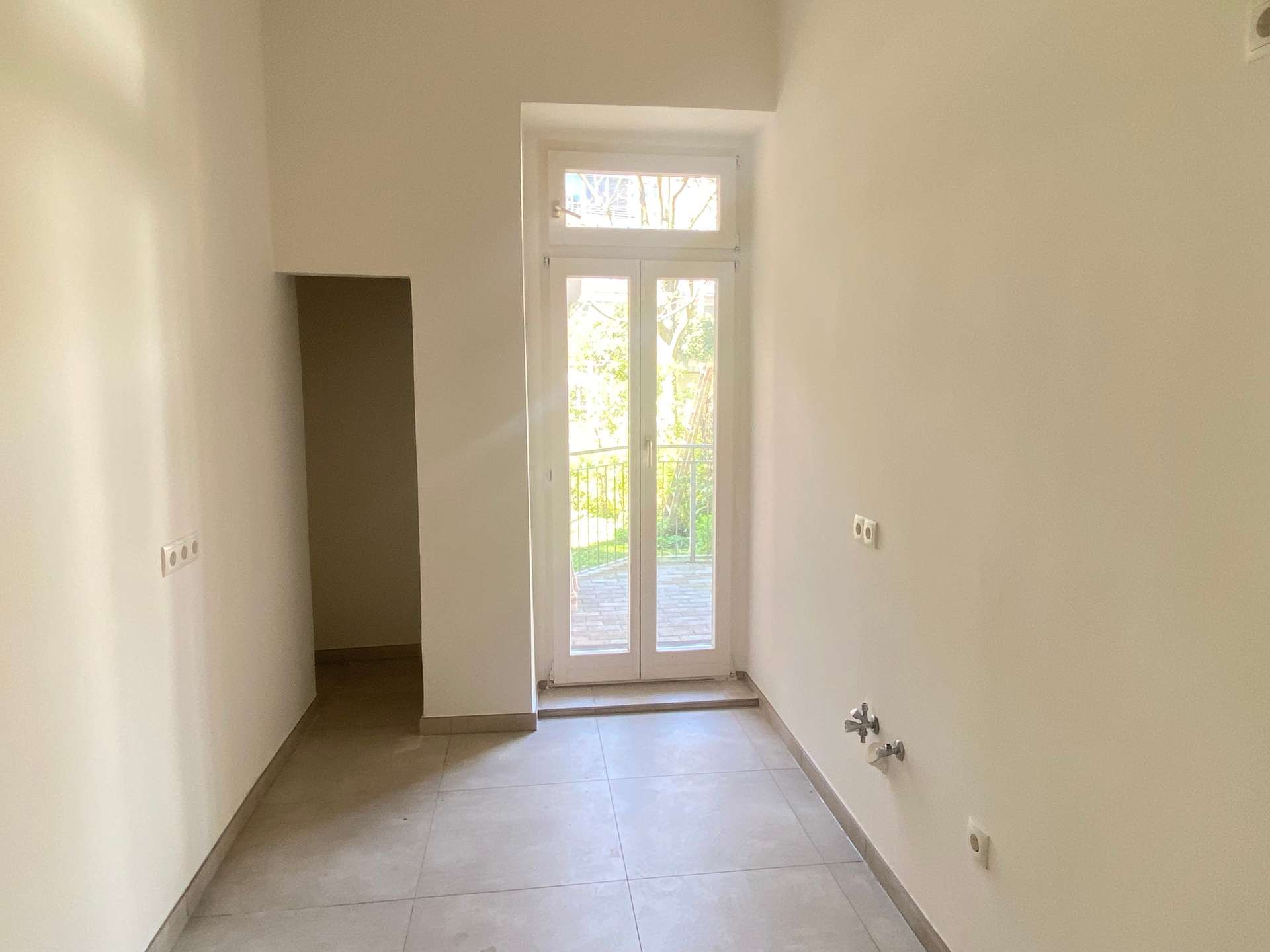 Narrow hallway with pale walls and tiled floor, ending at glass French doors that open to an outdoor area.