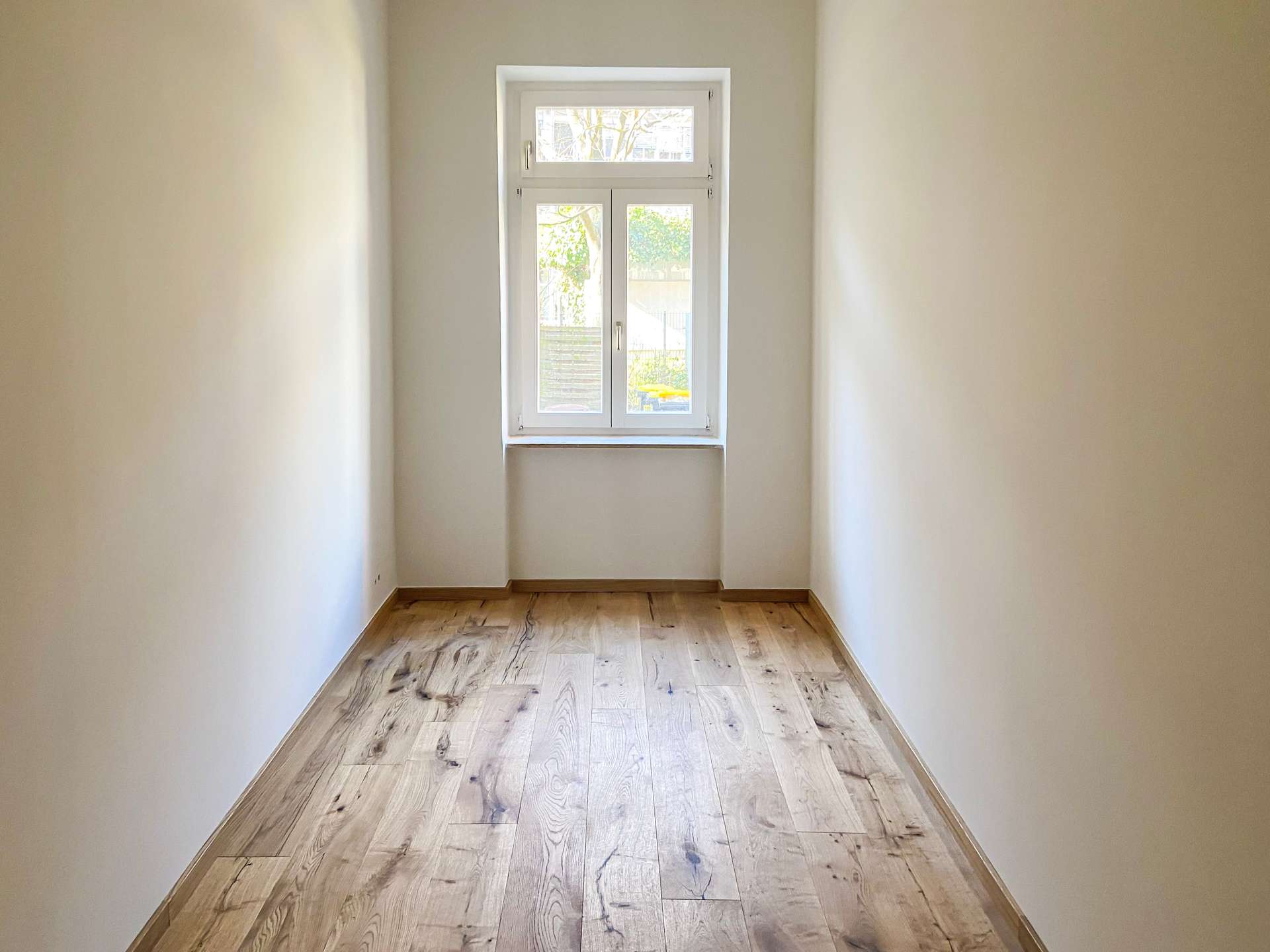 Narrow empty hallway with light wooden floor and white walls, window at far end bringing in sunlight.