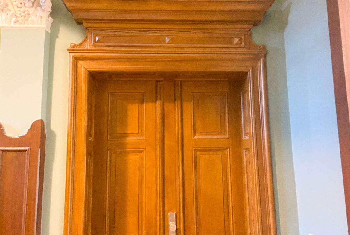 Tall wooden door with raised panels and decorative molding, set in a sage-green wall.