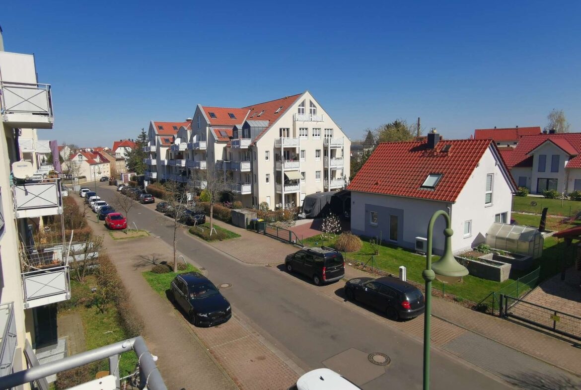 Sunny residential street with white apartment buildings and red-tiled roofs, parked cars along the curb under a clear blue sky.