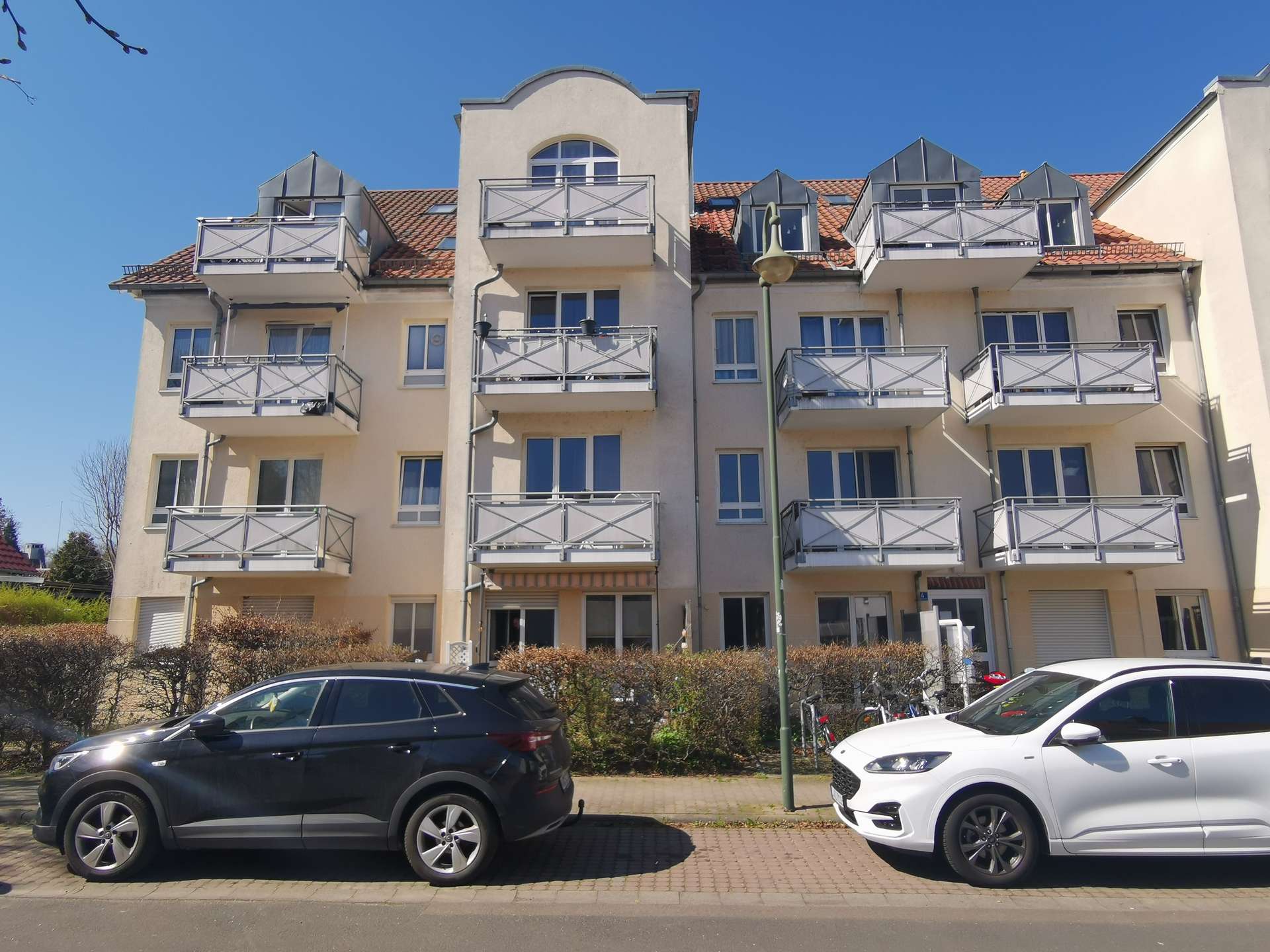 Residential building with multiple balconies and a blue sky, cars parked along the street in front.