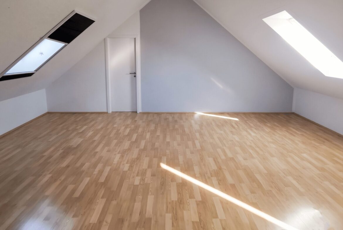 Empty attic room with sloped white walls, a white door, and light wood flooring under a peaked ceiling with skylights on both sides.