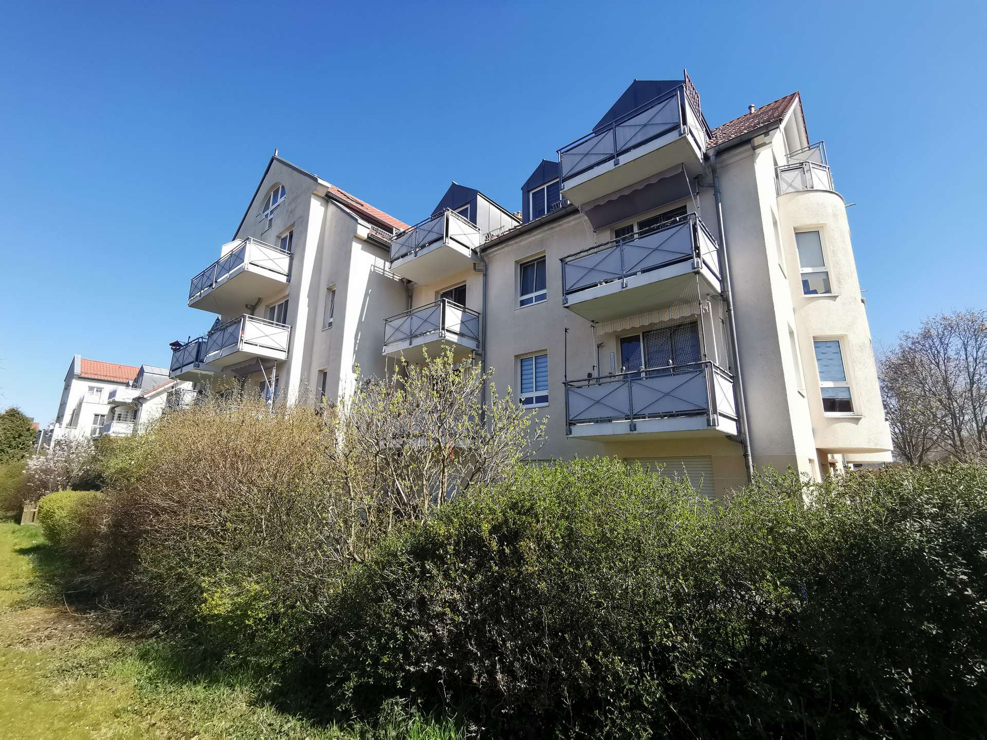 Apartment building with multiple balconies set against a clear blue sky, surrounded by shrubs and greenery in the foreground.