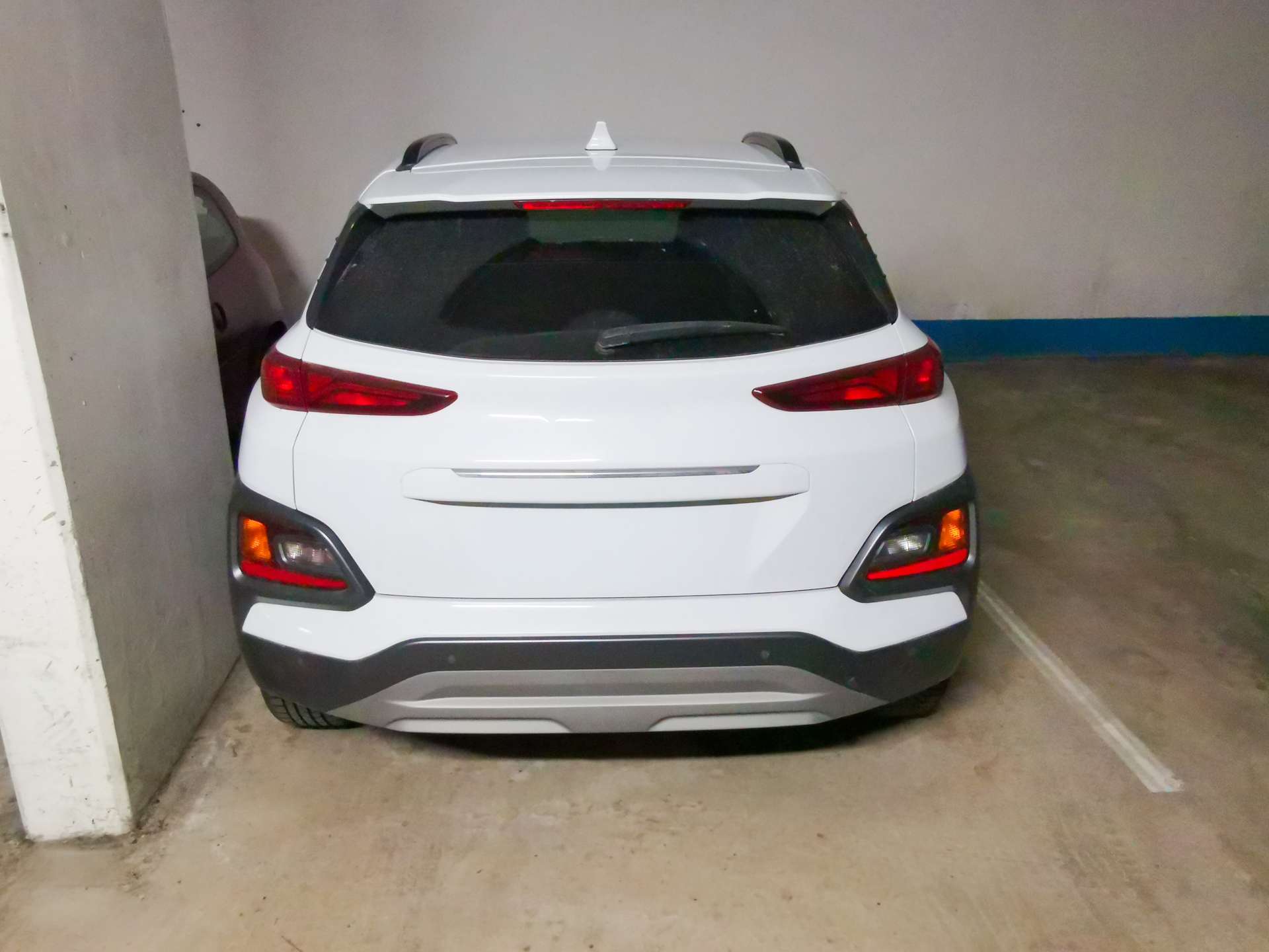 White SUV viewed from the rear in a dim parking garage, between a concrete pillar and a wall with a blue baseboard.
