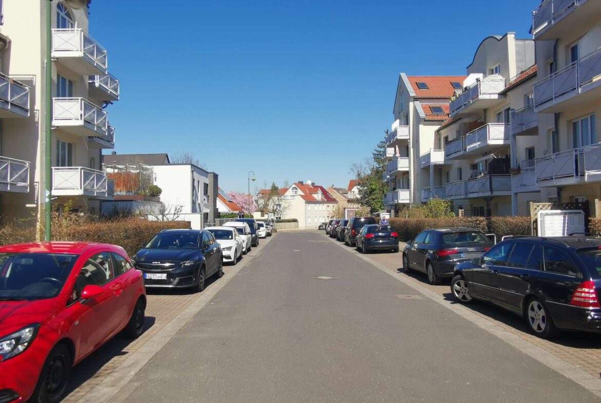 Street lined with modern apartment buildings on both sides and rows of parked cars under a clear blue sky with no pedestrians in sight.