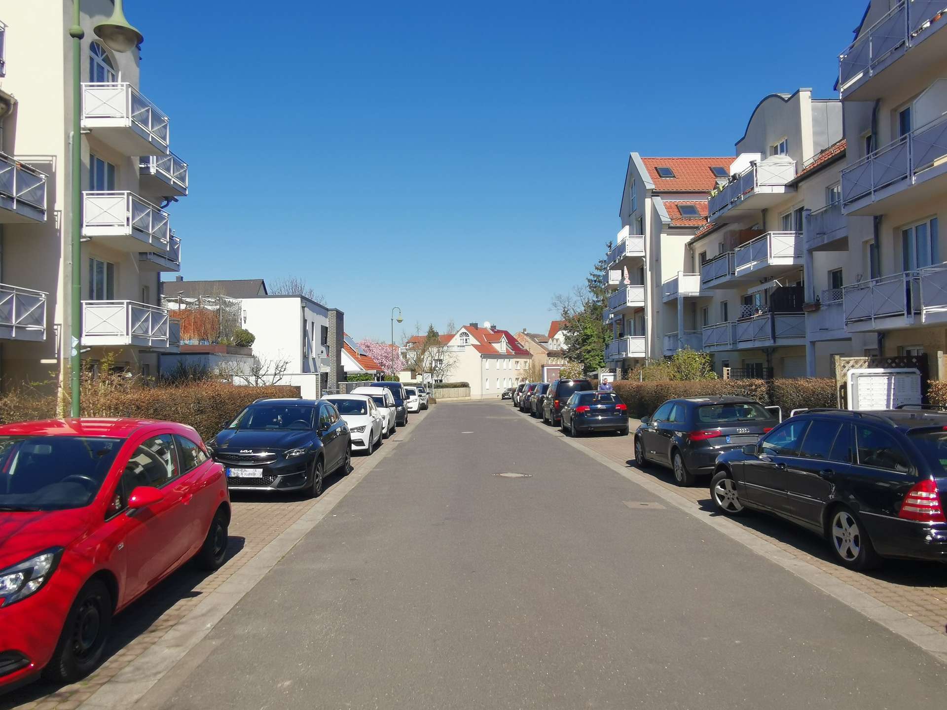 Street lined with modern apartment buildings on both sides and rows of parked cars under a clear blue sky with no pedestrians in sight.