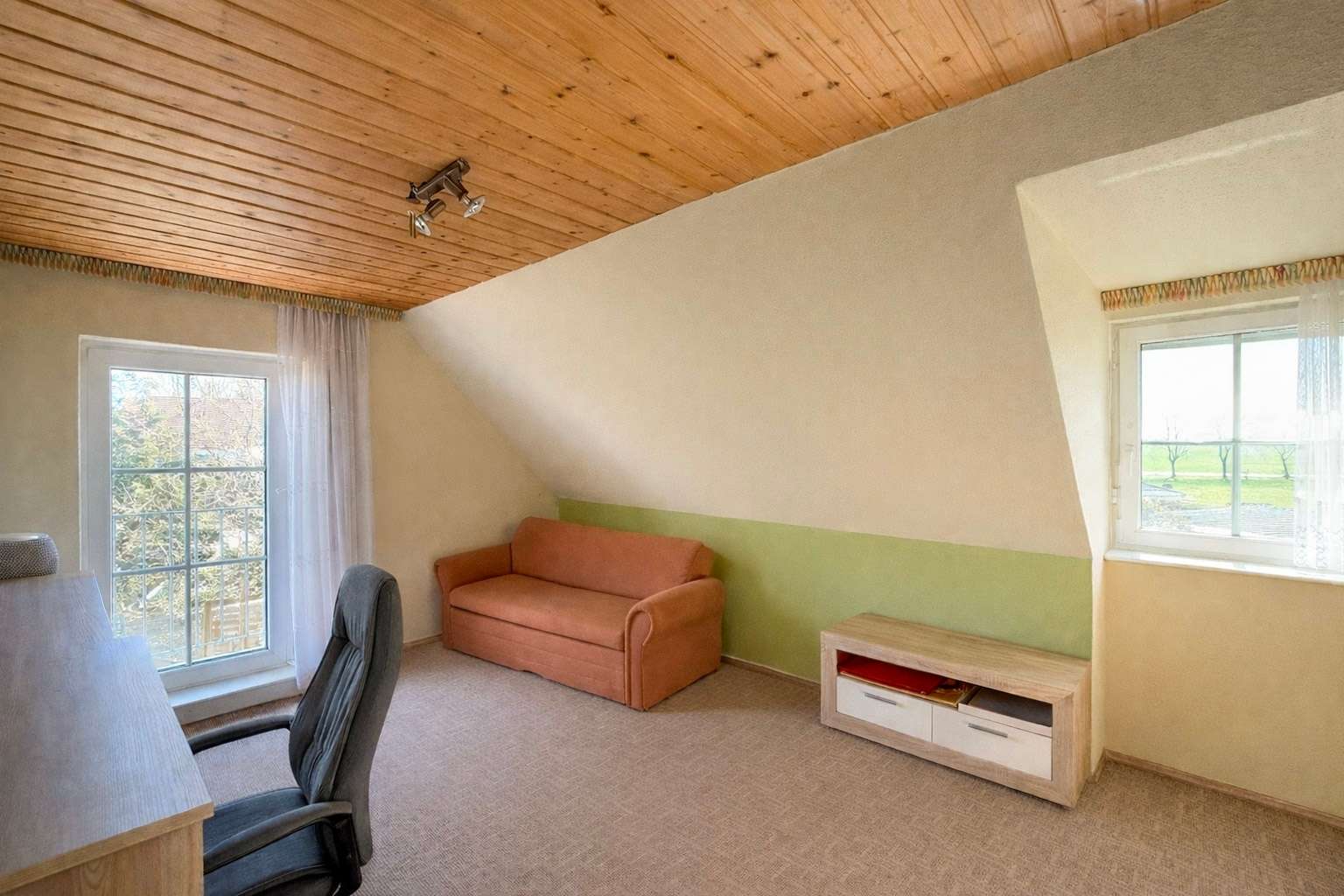 Cozy attic living room with a wood-paneled ceiling, peach sofa, desk, and a chair by a glass door with sheer curtains.