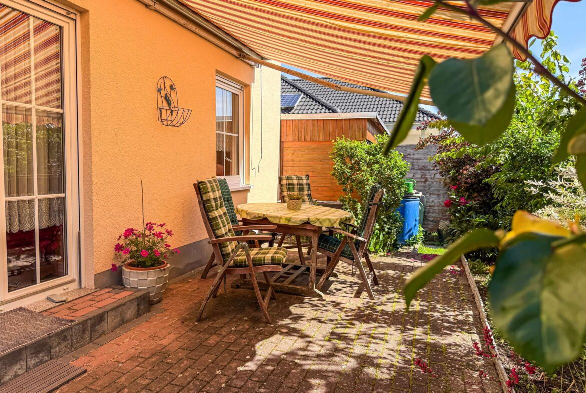 Outdoor patio with a round wooden table and plaid chairs under a striped awning, against an orange house wall with potted flowers nearby.