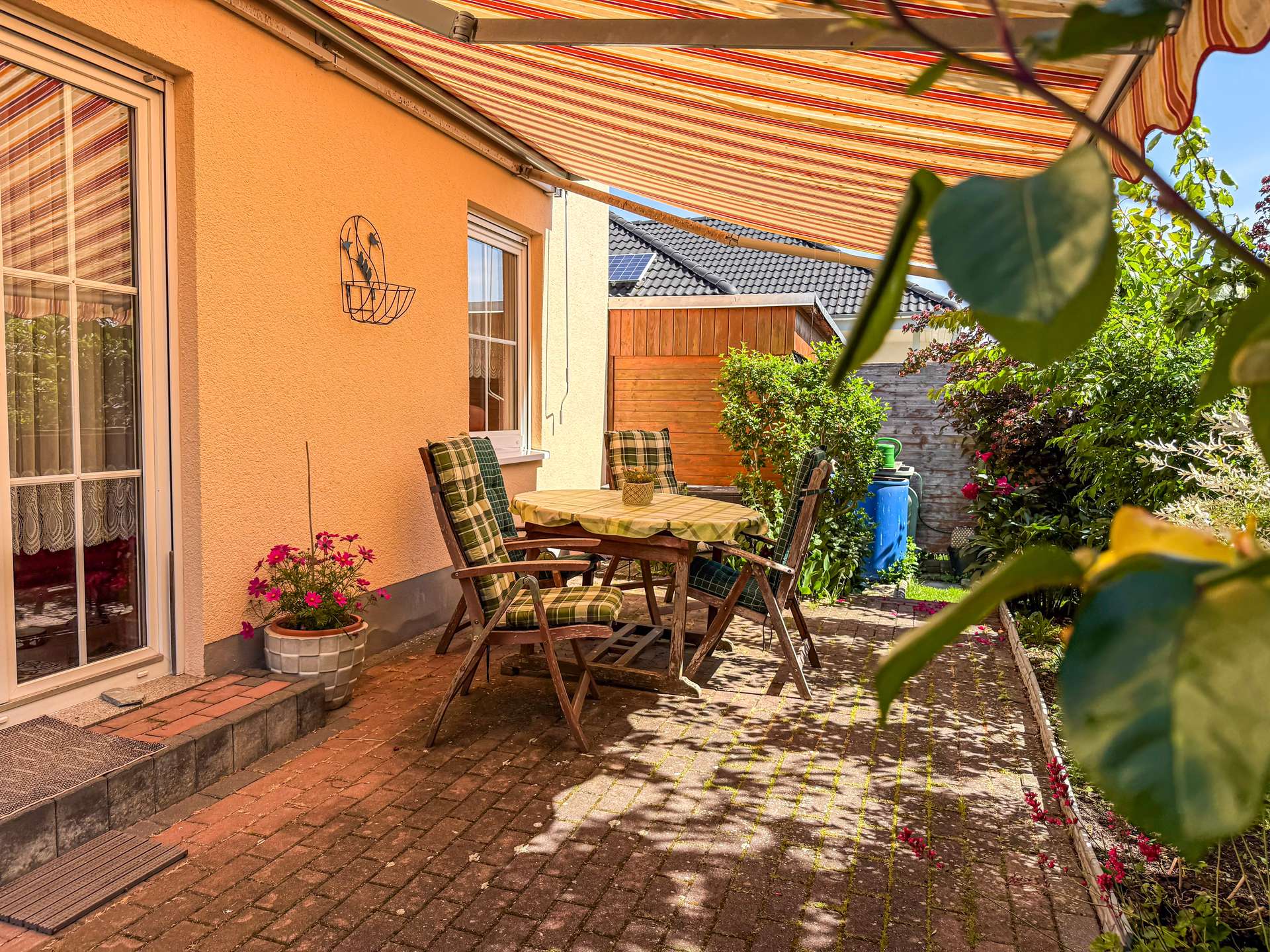 Outdoor patio with a round wooden table and plaid chairs under a striped awning, against an orange house wall with potted flowers nearby.