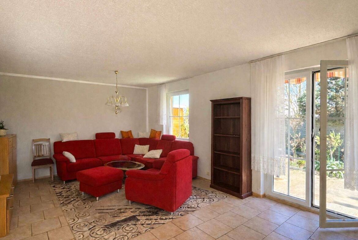 Bright living room with a red sectional sofa, ottoman, glass coffee table, and a tall wooden bookshelf by a glass door to the garden.