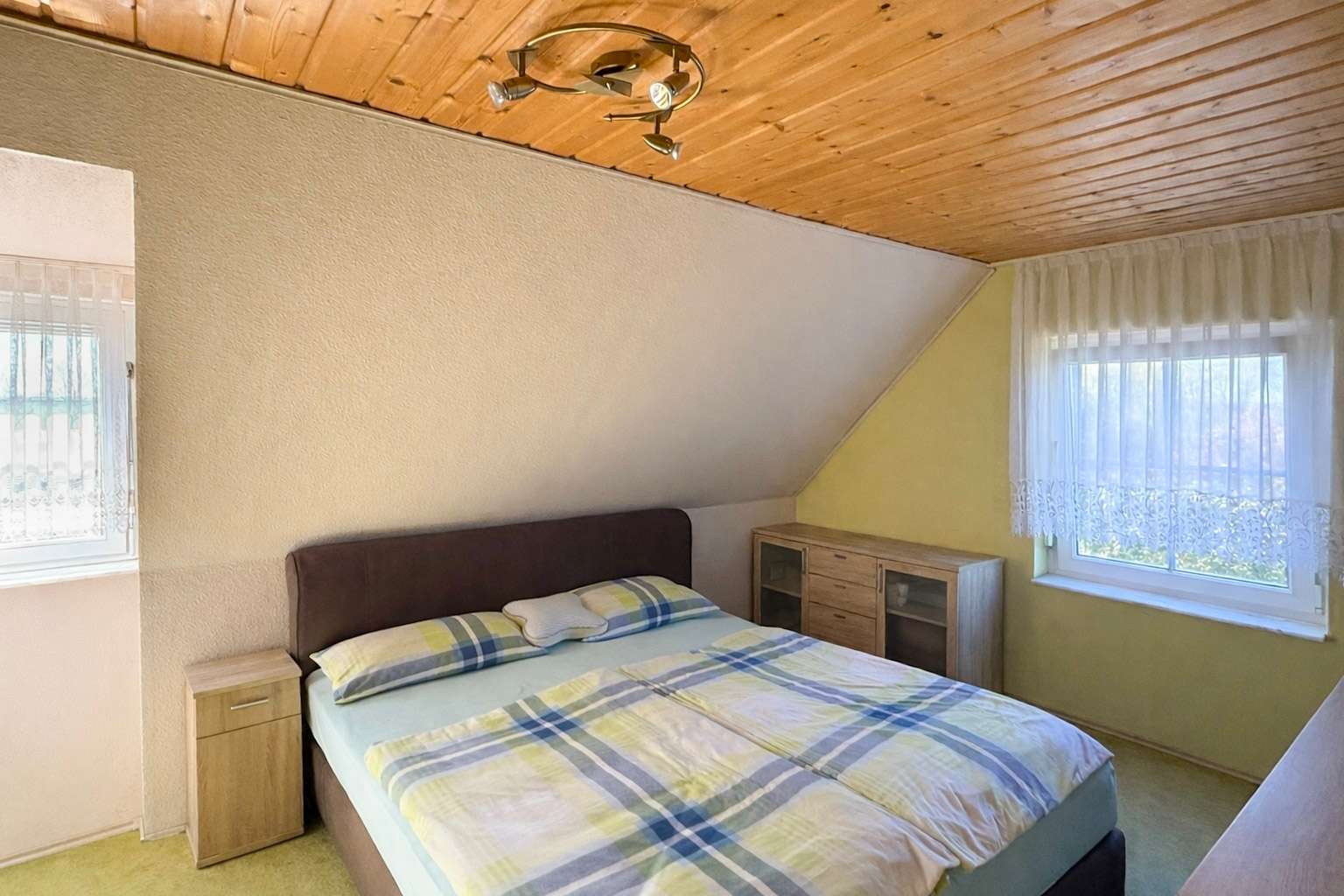 Cozy bedroom with a sloped wooden ceiling, a double bed with blue-yellow plaid bedding, and light wood nightstands and dresser.