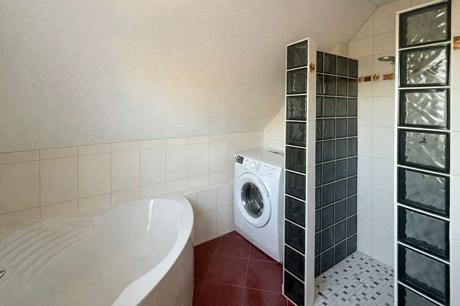 White corner bathtub in the foreground with a front-loading washing machine beside tiled walls and glass block partitions forming a shower area in a modern bathroom.