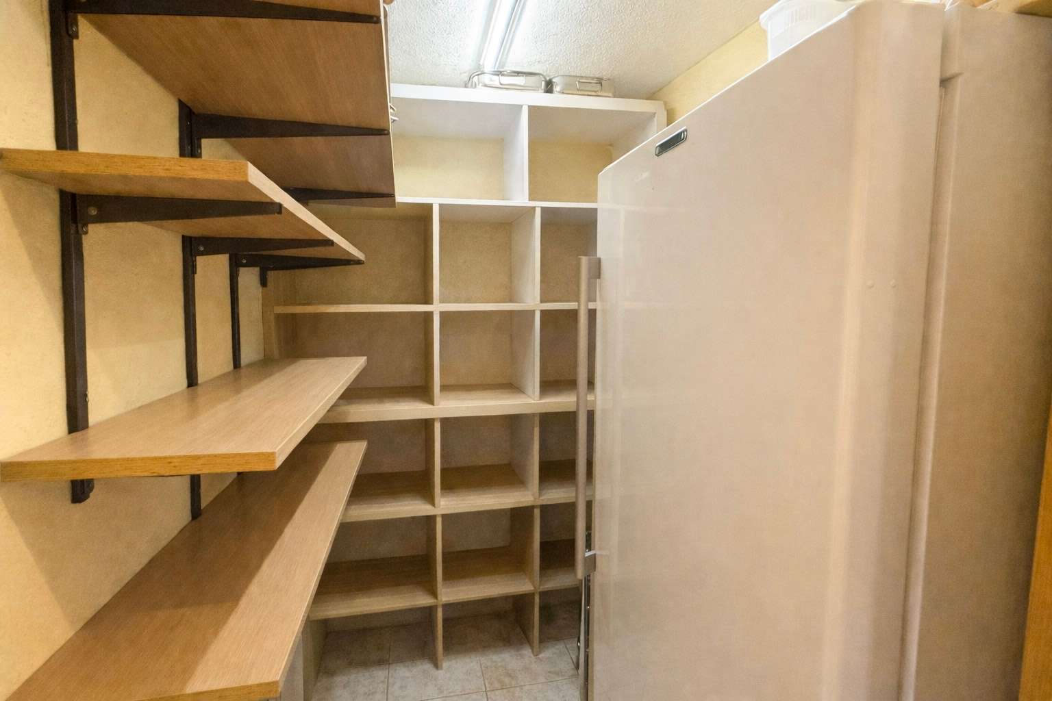 Narrow pantry with wooden shelves along the left wall, open cubby shelving in back, and a tall white fridge on the right.