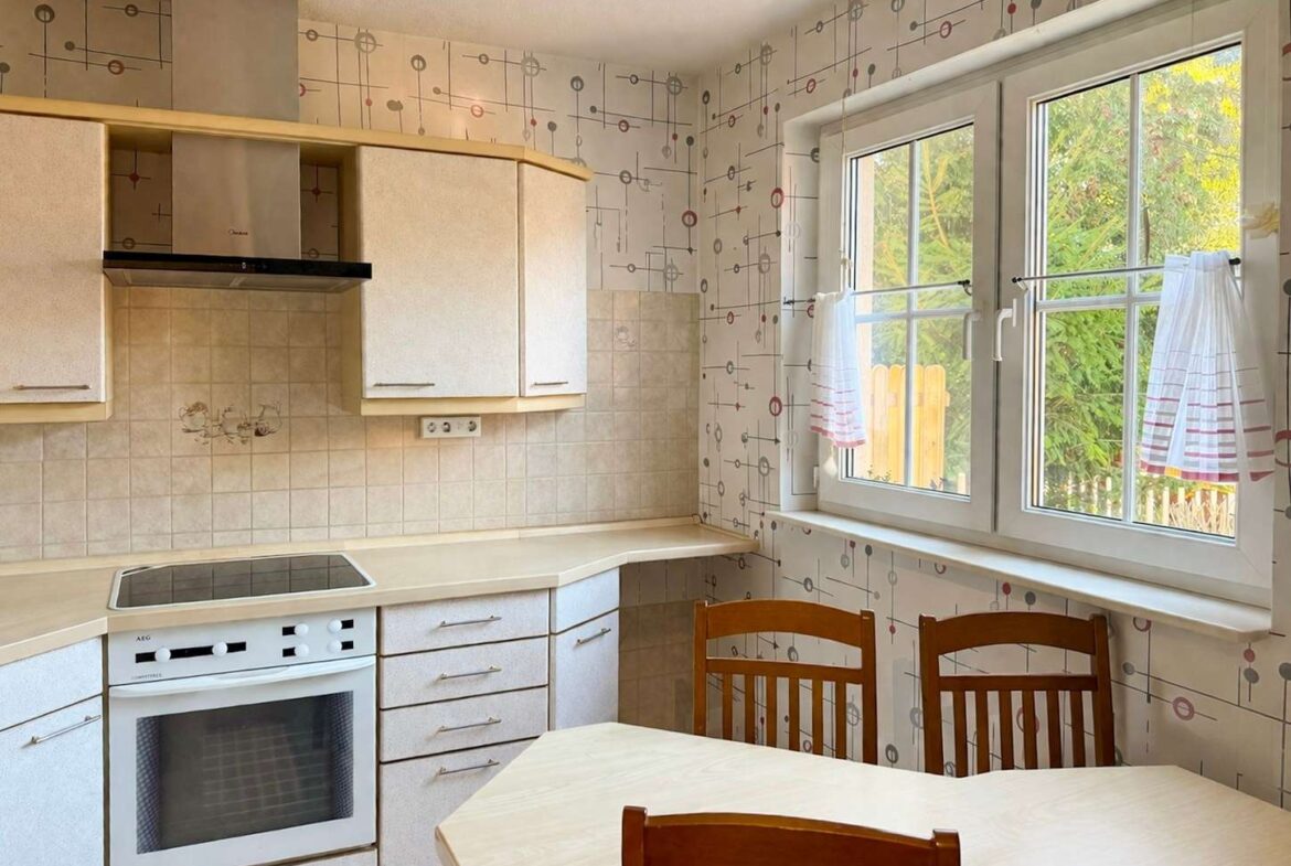 Cozy retro kitchen with beige cabinets, built-in oven and electric stove, tiled backsplash, and a wooden dining table with chairs. Large window reveals greenery outside.