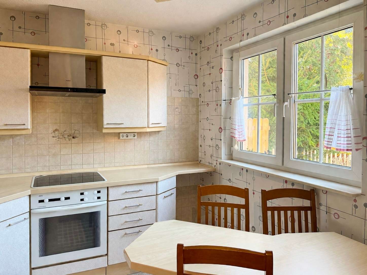 Cozy retro kitchen with beige cabinets, built-in oven and electric stove, tiled backsplash, and a wooden dining table with chairs. Large window reveals greenery outside.