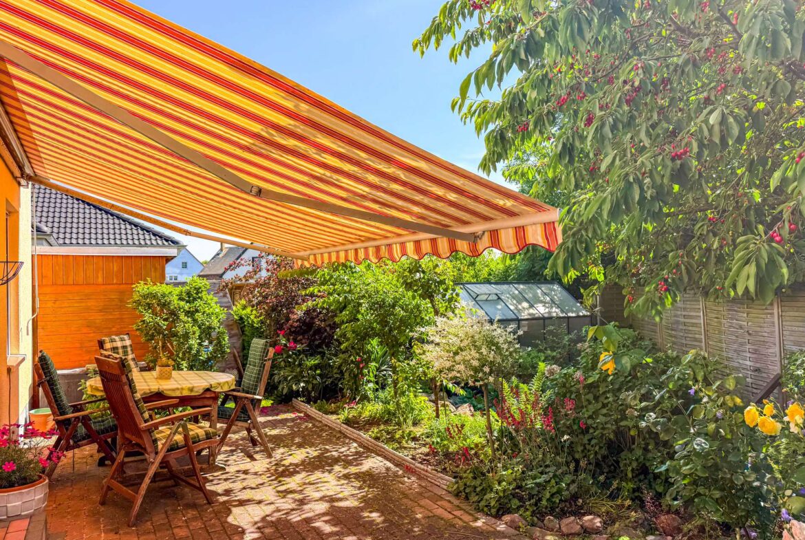Outdoor patio with wooden table and chairs under a bright orange striped awning, surrounded by lush garden plants.