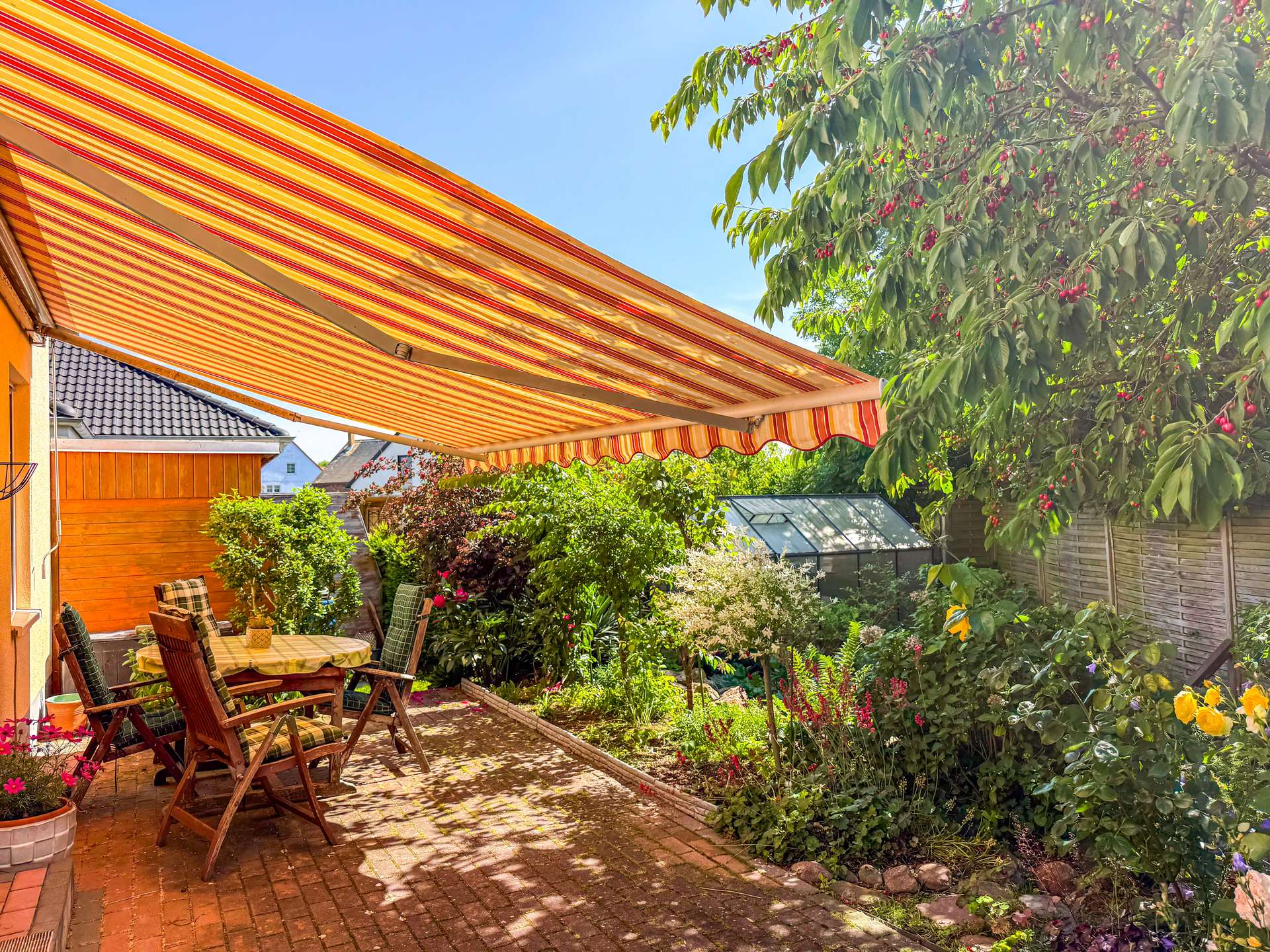 Outdoor patio with wooden table and chairs under a bright orange striped awning, surrounded by lush garden plants.