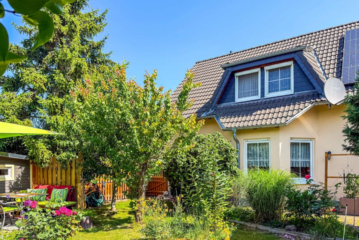 Sunny backyard with a two-story house, tiled roof, and satellite dish, surrounded by lush greenery and blooming flowers