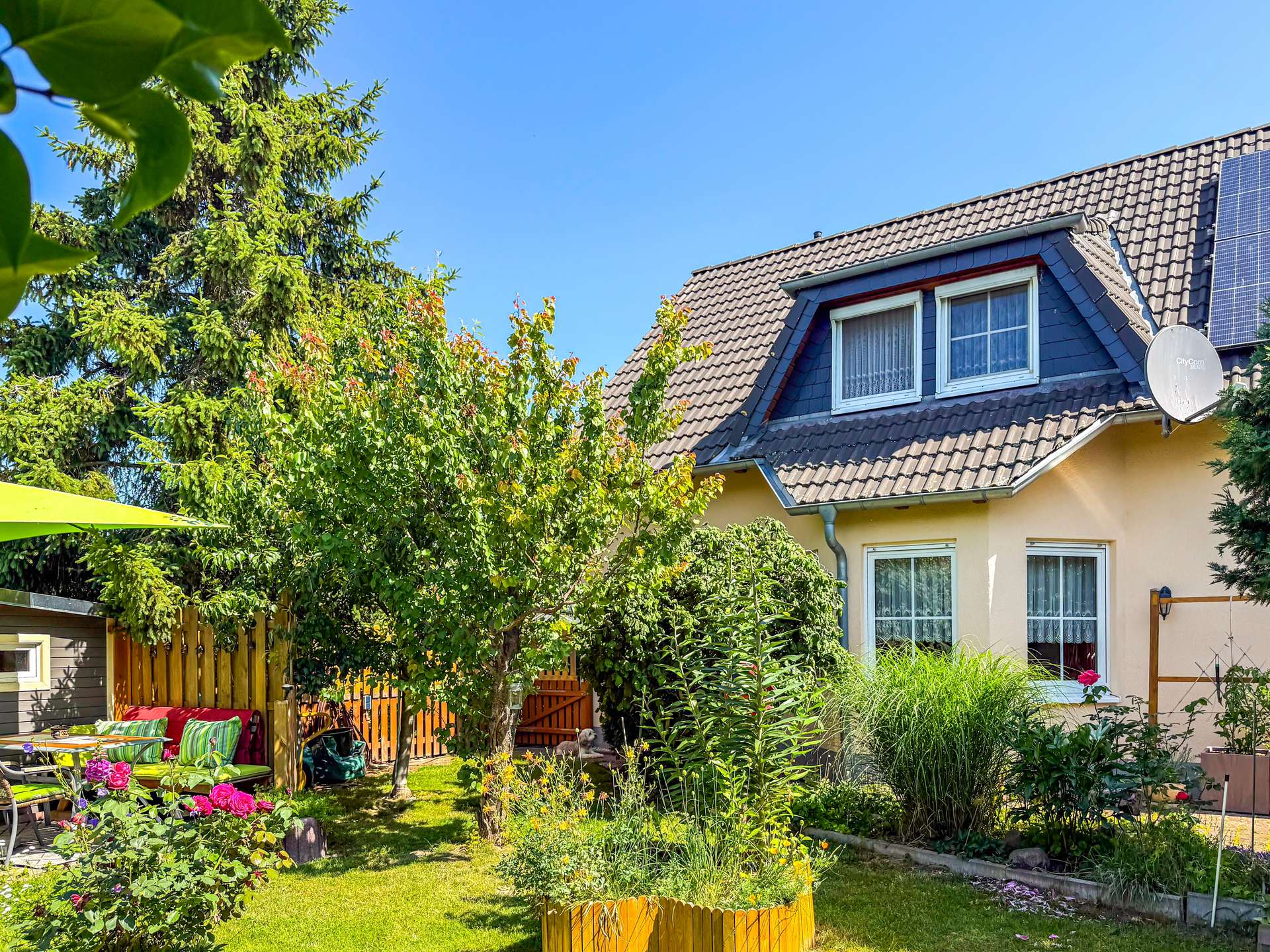 Sunny backyard with a two-story house, tiled roof, and satellite dish, surrounded by lush greenery and blooming flowers
