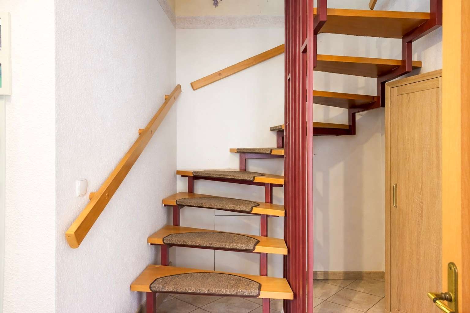 Interior staircase with open risers, orange wooden steps, and a red metal frame; a wooden handrail runs along the left wall, leading to a hallway and closet door on the right.