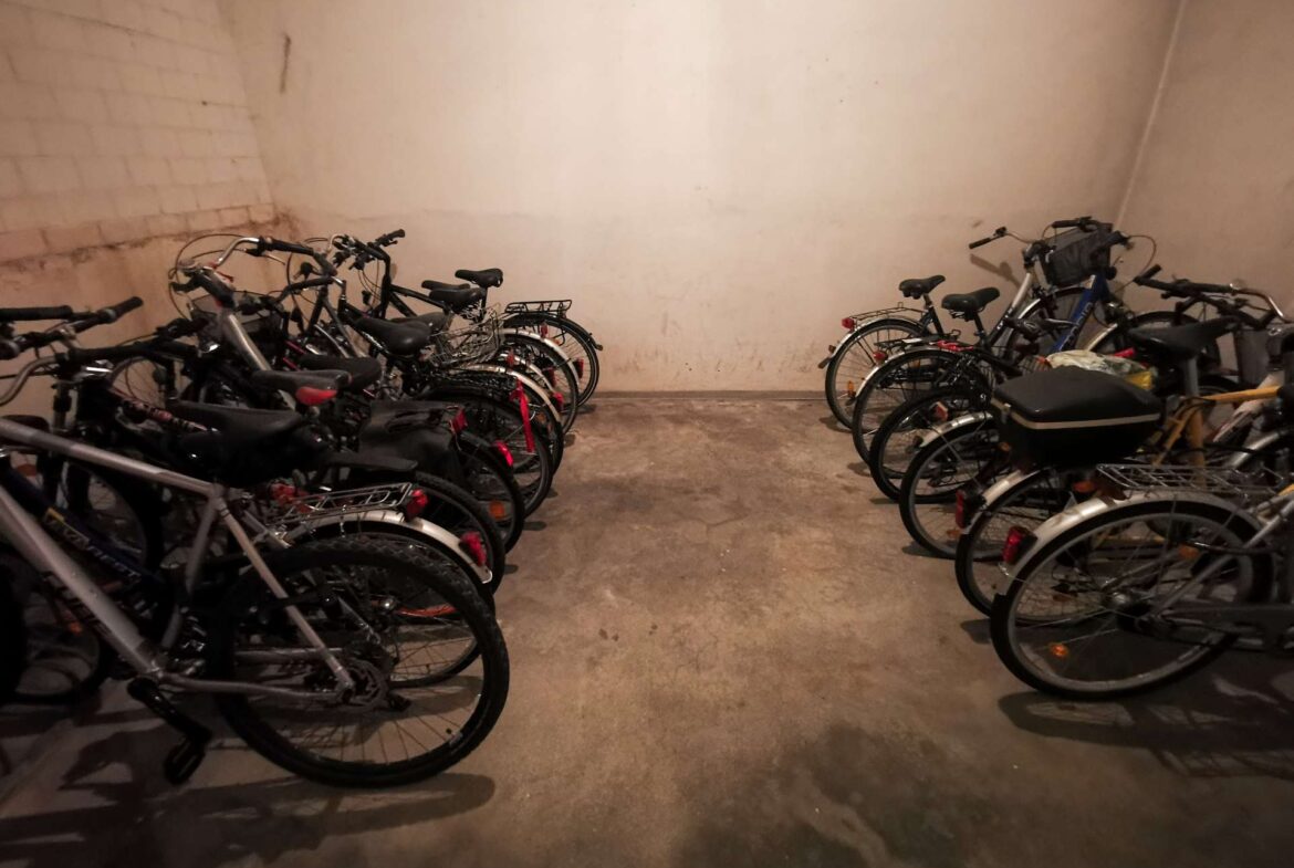 Bicycles parked along opposite walls in a dim underground bike storage area, concrete floor and bare walls visible