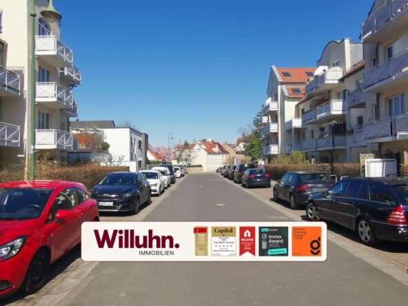 Residential street with parked cars on both sides and white apartment buildings; a large Willuhn Immobilien banner with award stickers in the foreground.