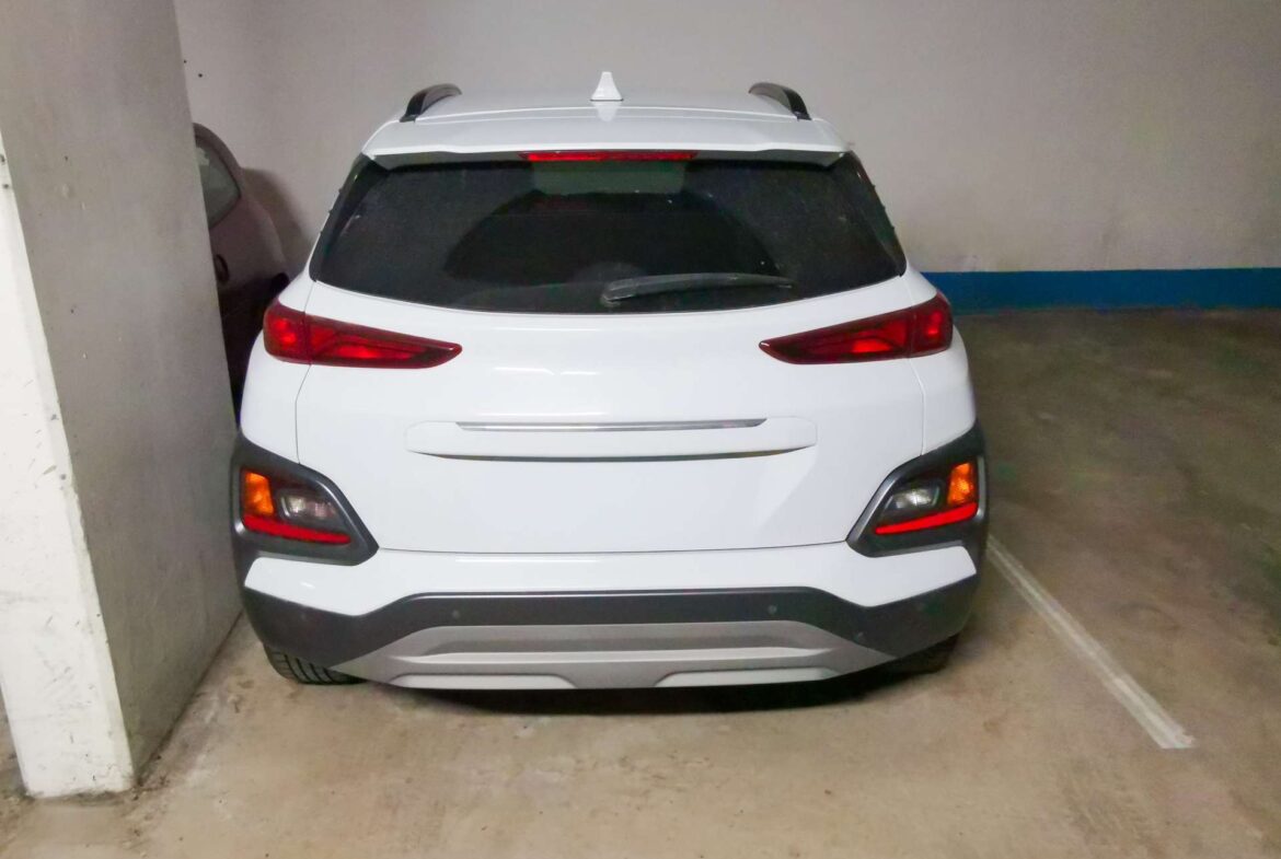White SUV shown from the rear parked in a dim indoor garage between a pillar and wall, roof rails visible.