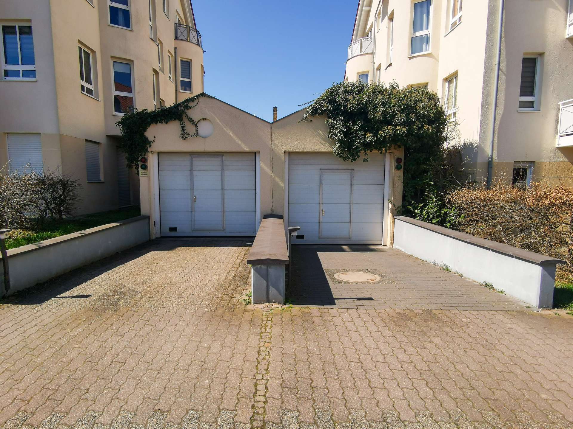 Beige residential buildings flank a driveway leading to two white garage doors with ivy climbing the walls under a clear blue sky.