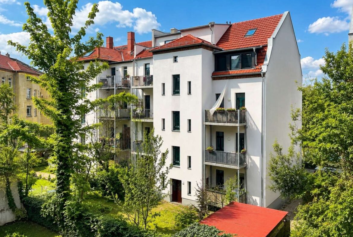 White, multi-story apartment building with red-tiled roof and balconies, surrounded by trees under a blue sky.