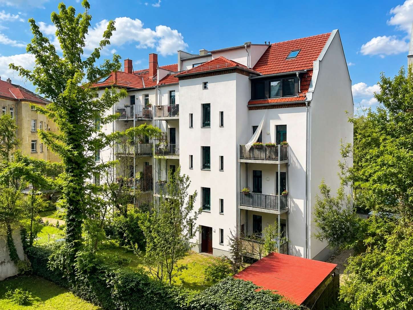 White, multi-story apartment building with red-tiled roof and balconies, surrounded by trees under a blue sky.
