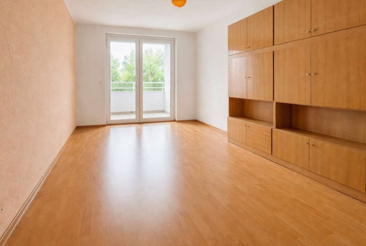 Empty living room with light wooden floor and built-in cabinets on the right, leading to a balcony through glass doors.