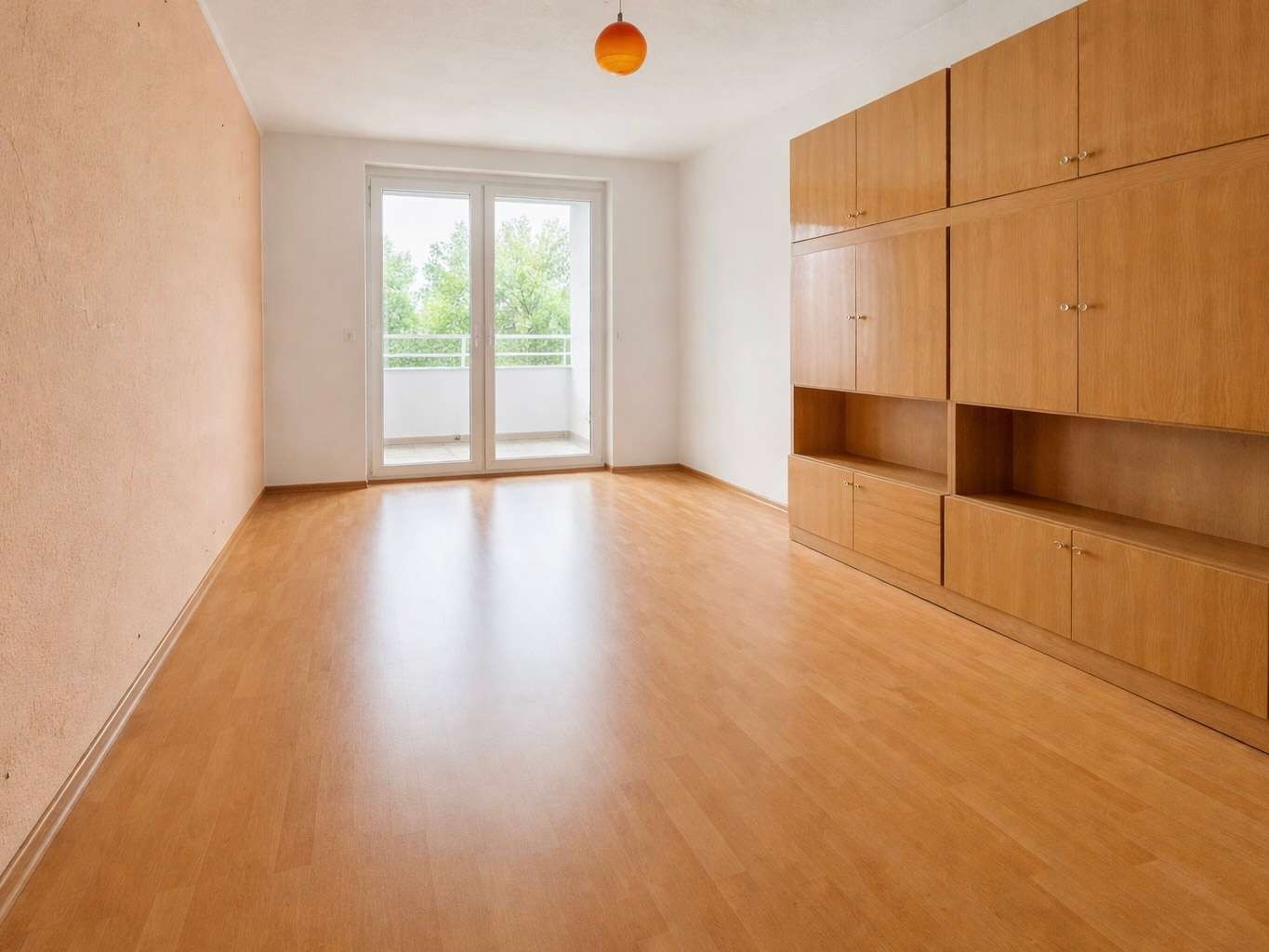 Empty living room with light wooden floor and built-in cabinets on the right, leading to a balcony through glass doors.