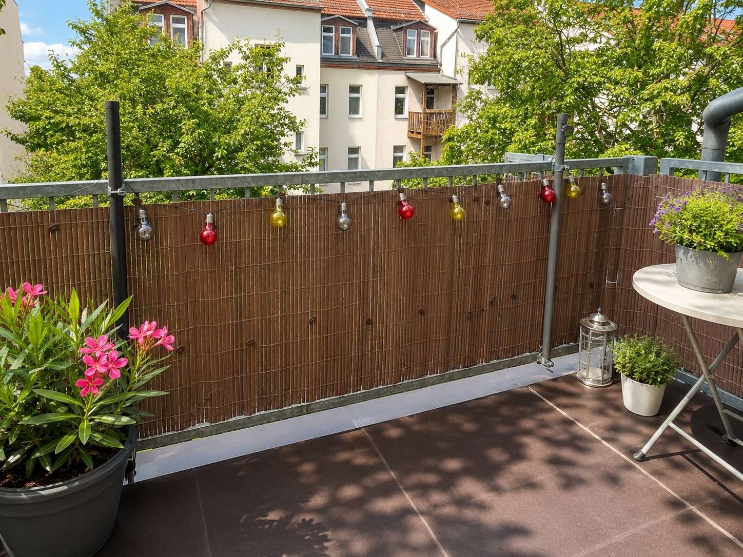 Cozy balcony with a brown bamboo screen, colorful bulb string lights, and potted plants including pink flowers; table with a lantern visible.