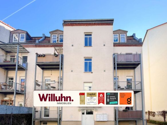 Apartment building with metal balconies and a large Willuhn Immobilien sign in front; blue sky above.
