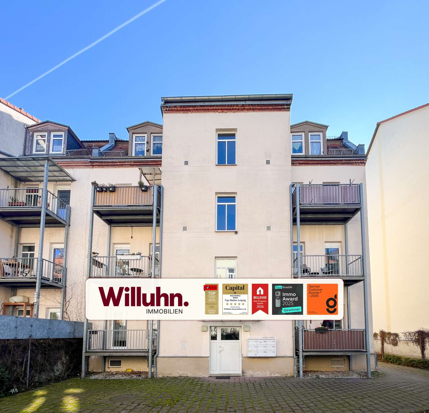Apartment building with metal balconies and a large Willuhn Immobilien sign in front; blue sky above.