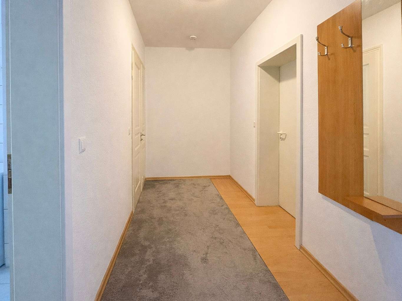 Wide hallway with white walls, beige doors, and a gray carpet runner along a light-wood floor; a wall mirror with hooks on the right.