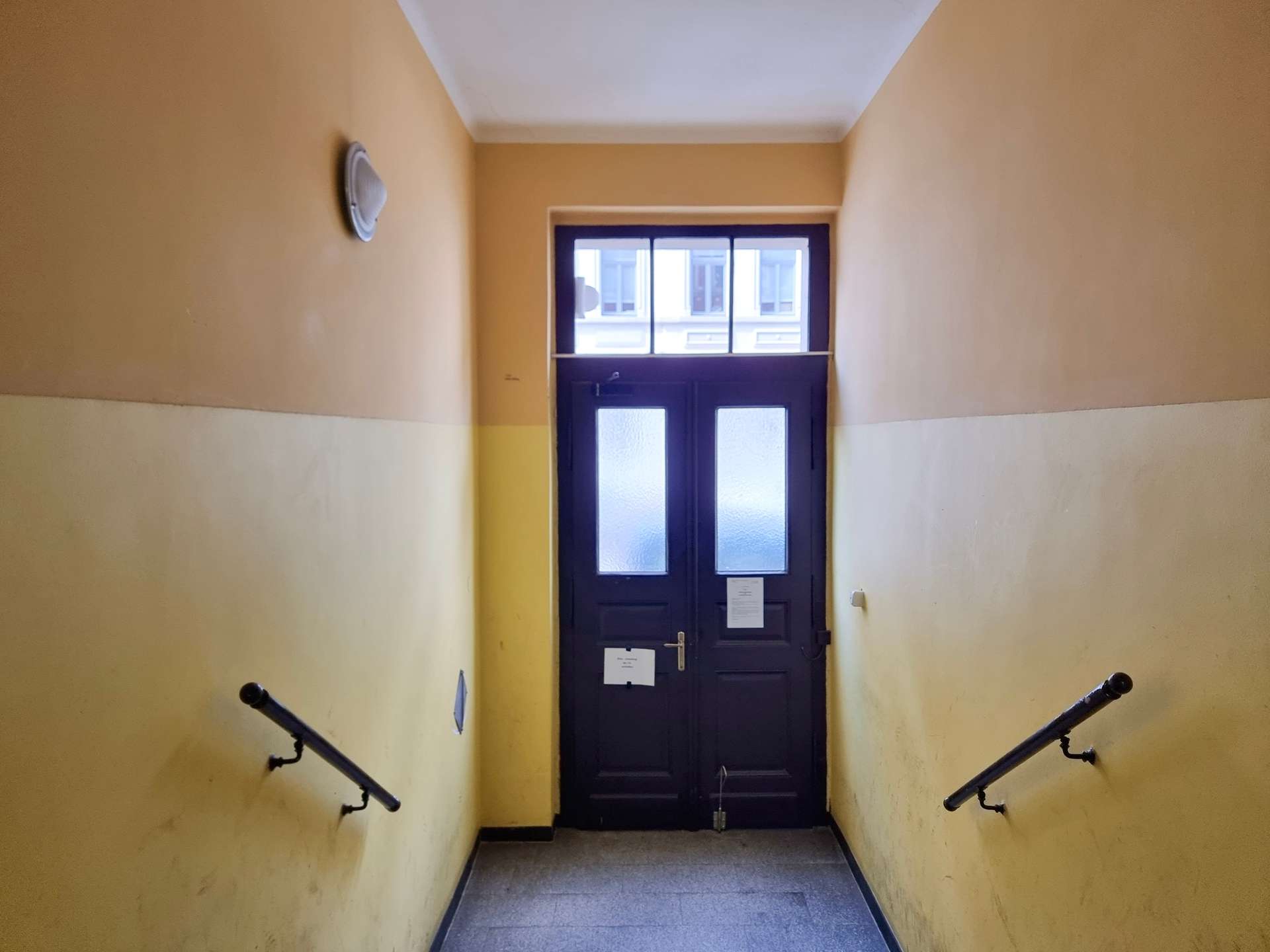 Narrow stairwell descending toward a dark door with frosted glass panels; yellow walls on both sides and metal handrails.